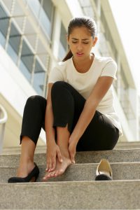 Portrait of latina woman walking on high heels and feeling pain, massaging feet with hand and sitting on stairs