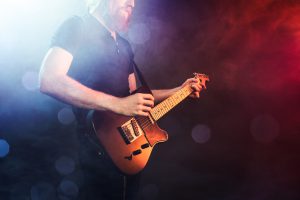 A fashionable young man with long hair and beard plays a black electric guitar at a live music venue, orange and blue stage lights illuminating the scene. Focus on the guitar. Horizontal image with copy space.