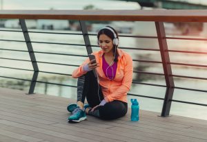 Young woman running in the city. She is sitting and taking break before she continue her daily routine. Sitting and using her smart phone. Texting. Water bottle is beside her.